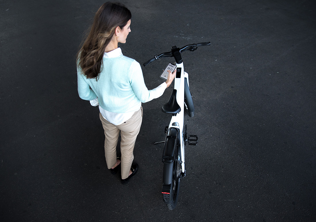 Mise à jour de l'application Stromer OMNI : femme vue de derrière avec un smartphone dans la main et un vélo électrique Stromer
